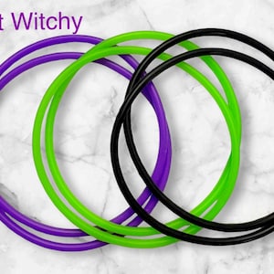 Może przedstawiać: Trzy okrągłe pierścienie w kolorze fioletowym, zielonym i czarnym są splecione na tle marmurowym. Tekst "Just Witchy" jest wyświetlany na górze po lewej stronie w kolorze fioletowym. Pierścienie wydają się być wykonane z elastycznego materiału.