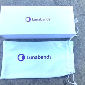 May include: A white rectangular box and a white drawstring bag. Both items have the purple text and logo "Lunabands". The box is open, revealing the bag. The background is a gray, textured surface.