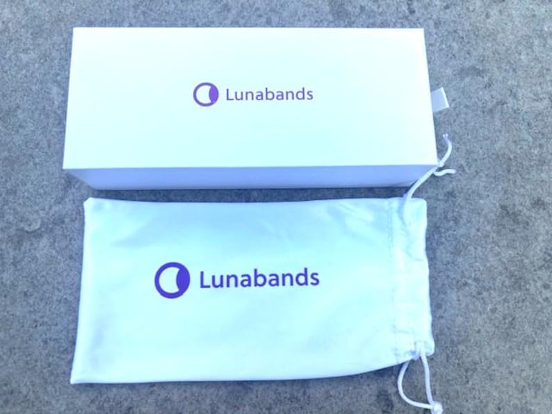 May include: A white rectangular box and a white drawstring bag. Both items have the purple text and logo "Lunabands". The box is open, revealing the bag. The background is a gray, textured surface.
