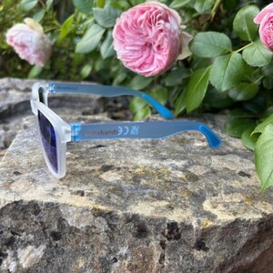 May include: White-framed sunglasses with blue arms and dark lenses rest on a textured stone surface. The sunglasses have the text "sunnebends" printed on the arm. Pink roses and green foliage are in the background.