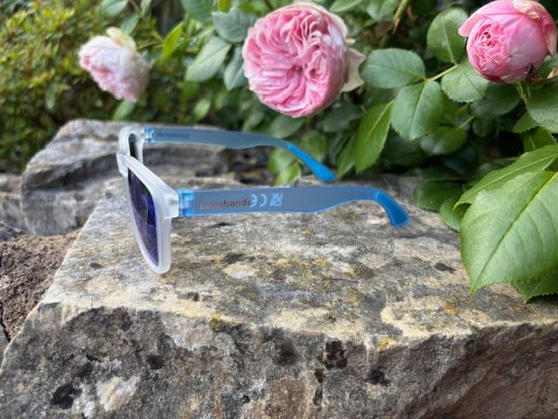 May include: White-framed sunglasses with blue arms and dark lenses rest on a textured stone surface. The sunglasses have the text "sunnebends" printed on the arm. Pink roses and green foliage are in the background.