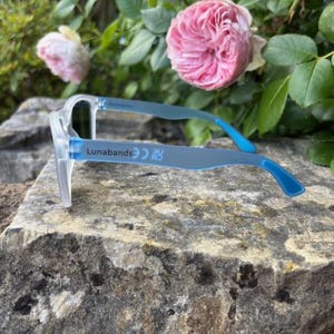 May include: Clear-framed eyeglasses with blue temples rest on a stone surface. The text "Lunabands" is printed on the temple. Pink roses and green foliage are in the background.