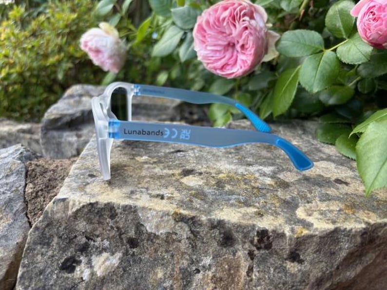 May include: Clear-framed eyeglasses with blue temples rest on a stone surface. The text "Lunabands" is printed on the temple. Pink roses and green foliage are in the background.