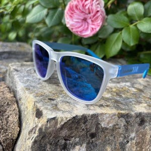 May include: White-framed sunglasses with blue lenses and blue arms rest on a textured stone surface. The sunglasses reflect a blue sky. Pink roses and green foliage are in the background.