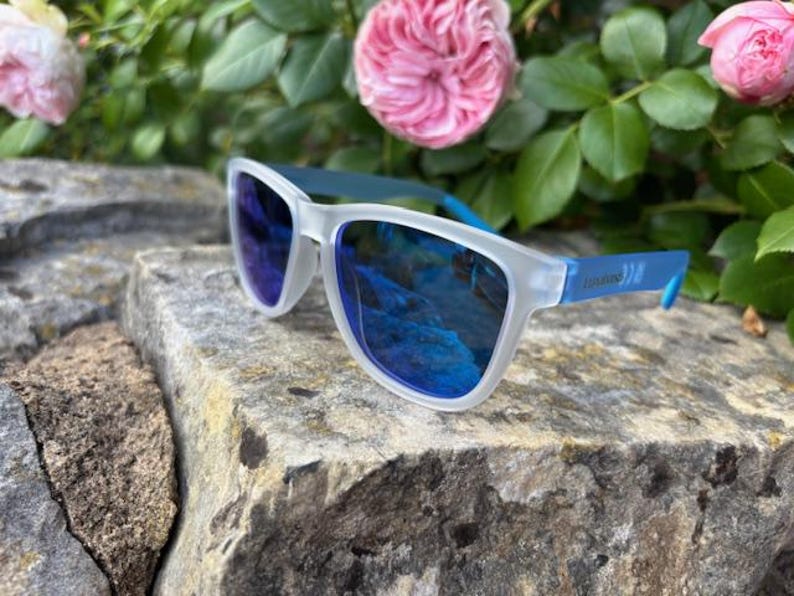 May include: White-framed sunglasses with blue lenses and blue arms rest on a textured stone surface. The sunglasses reflect a blue sky. Pink roses and green foliage are in the background.