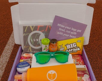 May include: An open box containing green sunglasses, snacks, and skincare products. The box includes an orange sunscreen tube, a purple card with text, and other small items. The box is placed on a running track.