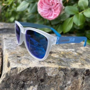 May include: Sunglasses with a white frame and blue arms, resting on a stone surface. The lenses are a reflective blue. The text "Lunatones" is printed on the arm. Pink roses and green foliage are in the background.