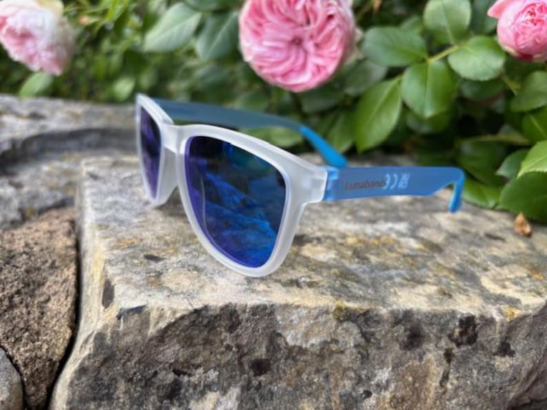May include: Sunglasses with a white frame and blue arms, resting on a stone surface. The lenses are a reflective blue. The text "Lunatones" is printed on the arm. Pink roses and green foliage are in the background.