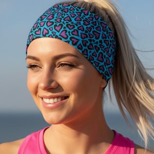 May include: A blue headband patterned with purple hearts. The headband is worn by a person with blonde hair and a pink top. The background shows a blurred view of the sky and water.