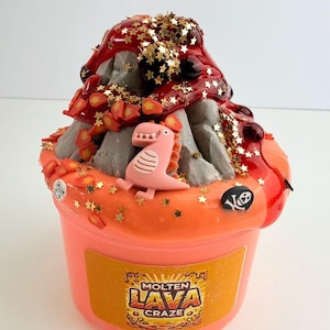 以下が含まれることがあります： 赤、オレンジ、金色のグリッターで飾られた火山型のトップを持つオレンジ色のスライム。火山にはピンクの恐竜が乗っており、頭蓋骨のチャームが付いています。スライムは、透明なプラスチック容器に入っていて、黄色いラベルには「Molten Lava Craze」と書かれています。