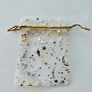 May include: A white sheer drawstring bag with a gold trim and a pattern of gold stars and crescent moons.