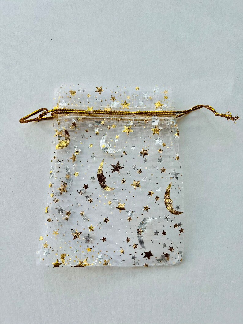 May include: A white sheer drawstring bag with a gold trim and a pattern of gold stars and crescent moons.