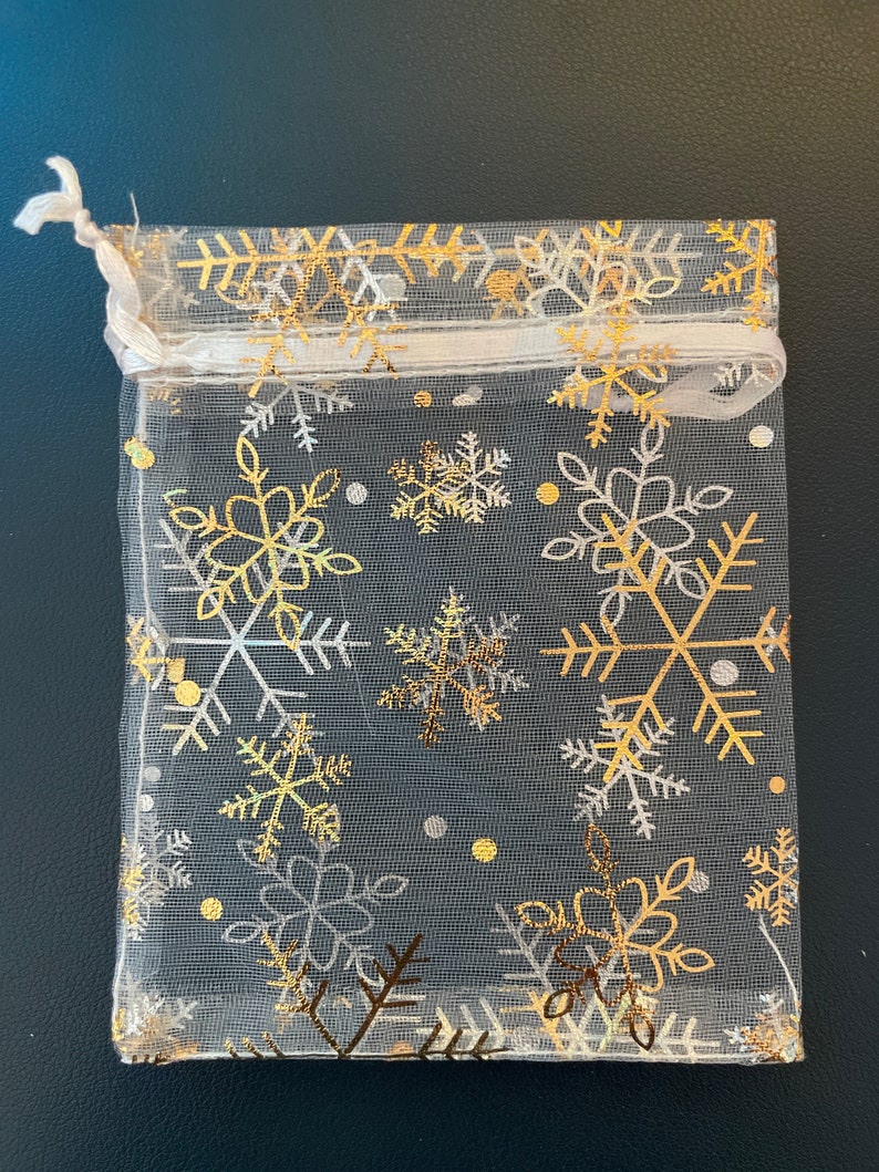 May include: A white sheer gift bag with a gold and white snowflake pattern. The bag has a drawstring closure.