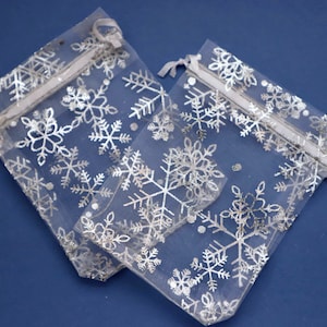 May include: Two white sheer organza gift bags with silver snowflake patterns. The bags have white drawstrings.