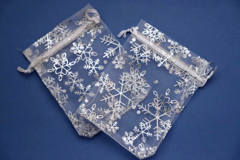 May include: Two white sheer organza gift bags with silver snowflake patterns. The bags have white drawstrings.