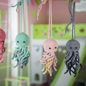 May include: Five crocheted jellyfish hanging from the ceiling. The jellyfish are pink, green, light pink, gray, and brown. They are all made with yarn and have small black eyes.