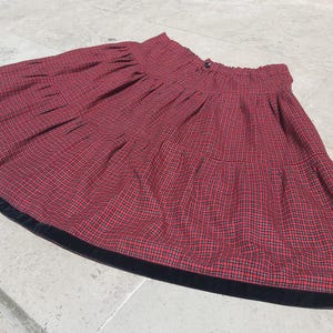 Pode incluir: Saia xadrez vermelha e preta com acabamento em veludo preto. A saia tem cintura franzida e fecho de botão. O tecido tem um visual clássico e vintage.