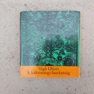 May include: A book cover featuring a green and black ornate pattern. The bottom section is orange and displays the text "Végh Olivér A kalotaszegei fazekasság". The book rests on a grey surface.