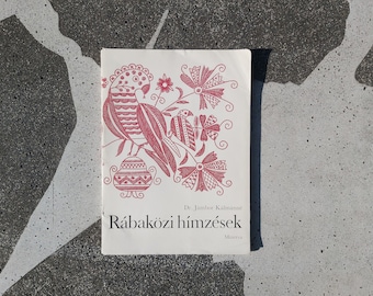 May include: A white book with a red bird and floral design on the cover. The title is "Rábaközi hímzések" by Dr. Jámbor Kálmánné. The book is published by Minerva.