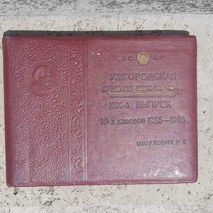 May include: Maroon-colored book with embossed portrait and text. The text reads "Uzhgorod Secondary School No. 1" and "10-X Classes 1965-1966." A gold emblem is at the top. The book is likely a yearbook or commemorative item.