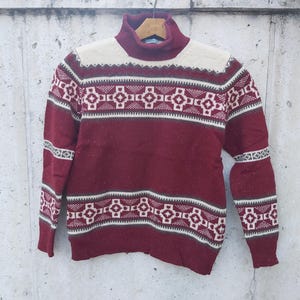 May include: A burgundy turtleneck sweater with a geometric pattern in white, brown, and cream. The sweater features a high neck and long sleeves, with the pattern repeating across the chest, sleeves, and upper back. The sweater is made of a soft, possibly wool, material.