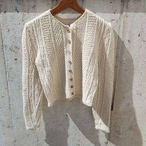 Könnte beinhalten: Ein weißer Strickcardigan mit Zopfmuster und Knopfleiste.