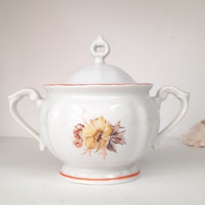 May include: White porcelain sugar bowl with a lid and two handles. The bowl has a floral design with yellow, brown, and orange flowers. The lid has a small knob on top.