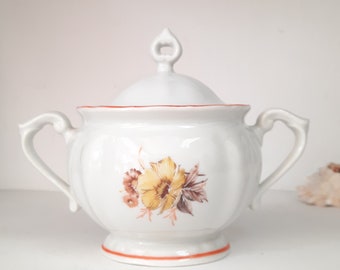 Peut inclure: Sucrier en porcelaine blanche avec un couvercle et deux anses. Le bol présente un motif floral avec des fleurs jaunes, brunes et orange. Le couvercle a un petit bouton sur le dessus.