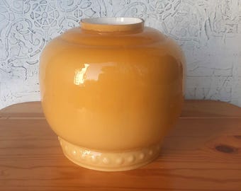 May include: A glossy, amber-colored decorative vase with a rounded body and a slightly flared base. The vase has a white interior and sits on a wooden surface. The base features a decorative, raised pattern.