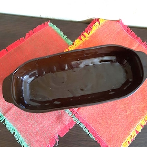 May include: A dark brown, oval-shaped ceramic baking dish with handles. The dish is empty and sits on a red and orange fringed placemat. The dish has a glossy finish and is likely used for cooking and serving food.