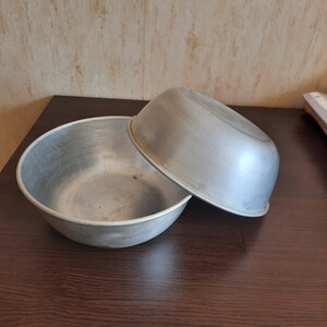 May include: Two silver metal bowls, one nested inside the other. The bowls are smooth and have a slightly worn appearance.