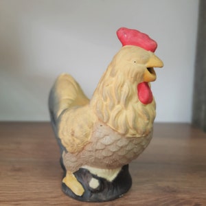 Peut inclure: Une figurine de coq en c&eacute;ramique avec un corps jaune, des plumes de queue noires et une cr&ecirc;te et des barbillons rouges. Le coq est debout sur une base brune.