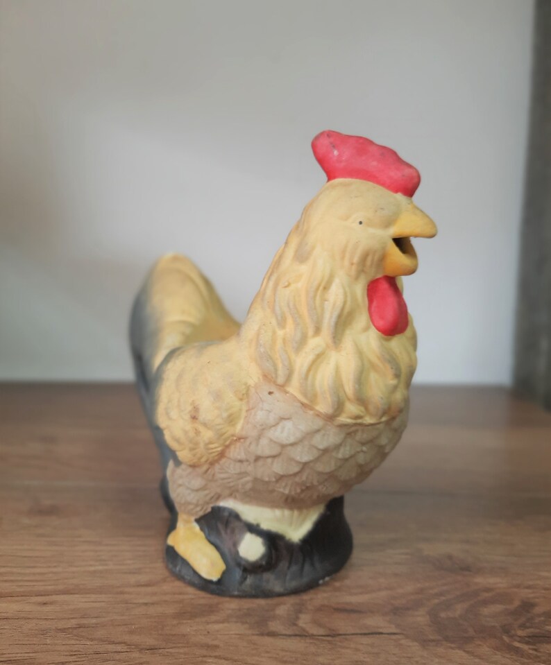 Peut inclure: Une figurine de coq en c&eacute;ramique avec un corps jaune, des plumes de queue noires et une cr&ecirc;te et des barbillons rouges. Le coq est debout sur une base brune.