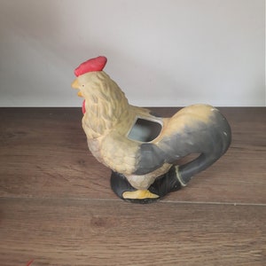 Peut inclure: Un pot de fleurs en c&eacute;ramique en forme de coq avec une cr&ecirc;te rouge et une queue noire et brune. Le coq est debout sur une surface en bois.