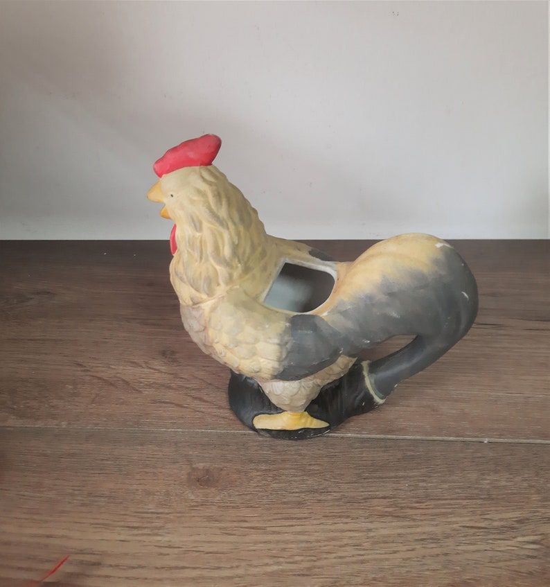 Peut inclure: Un pot de fleurs en c&eacute;ramique en forme de coq avec une cr&ecirc;te rouge et une queue noire et brune. Le coq est debout sur une surface en bois.