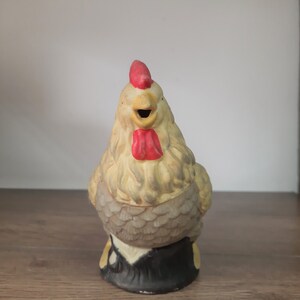 Peut inclure: Une figurine de coq en c&eacute;ramique avec une cr&ecirc;te et une barbe rouges, un bec jaune et des plumes grises et blanches. Le coq est debout sur une base noire.