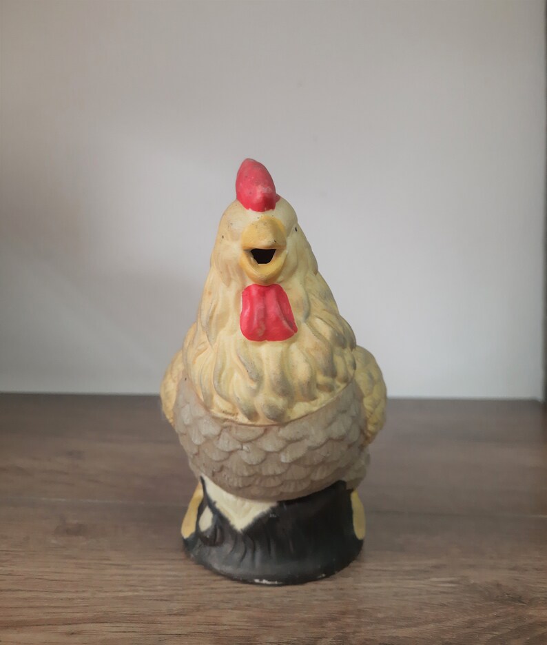 Peut inclure: Une figurine de coq en c&eacute;ramique avec une cr&ecirc;te et une barbe rouges, un bec jaune et des plumes grises et blanches. Le coq est debout sur une base noire.