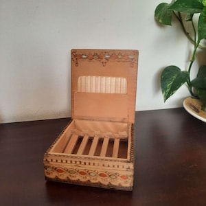 May include: Open wooden box with a hinged lid, featuring a decorative border with orange, blue, and green accents. The interior has a compartment with vertical dividers and a section for holding items. The box is made of light-colored wood.