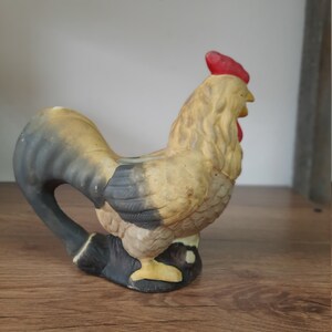 Peut inclure: Une figurine de coq en c&eacute;ramique avec un corps noir et brun, un bec jaune et une cr&ecirc;te rouge. Le coq est debout sur une surface en bois.
