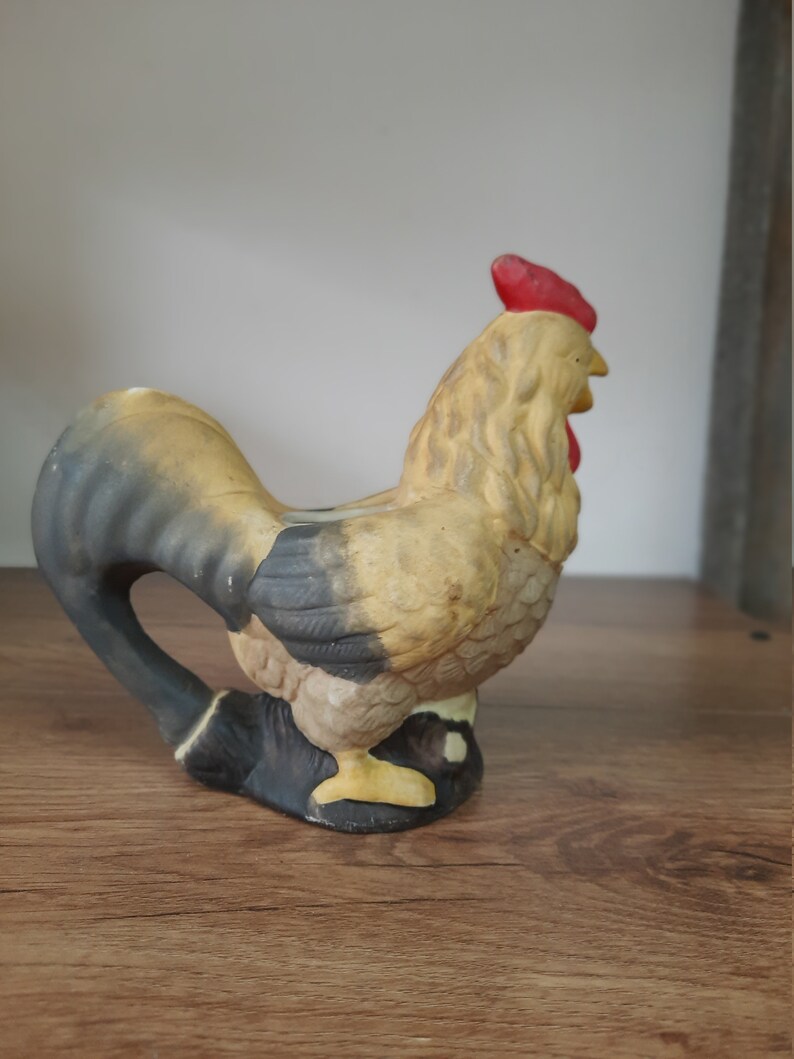 Peut inclure: Une figurine de coq en c&eacute;ramique avec un corps noir et brun, un bec jaune et une cr&ecirc;te rouge. Le coq est debout sur une surface en bois.