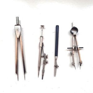 May include: Four silver compasses with different designs. Two compasses have a black handle and a silver needle. One compass has a silver handle and a silver needle. The fourth compass has a silver handle and a silver needle with a black ring.
