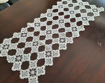 Peut inclure: Un chemin de table en crochet blanc avec un motif en losange. Le chemin de table est fait de points délicats et complexes et a un aspect de dentelle.