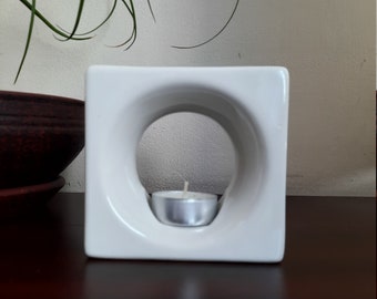 May include: White ceramic square candle warmer with a circular opening in the center. A small white tea light candle is placed inside the opening.