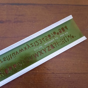 May include: A green plastic stencil ruler with a white border. The ruler has numbers, letters, and punctuation marks. The text on the ruler reads "1672 206 MEISSNER KG PEN 0.8".