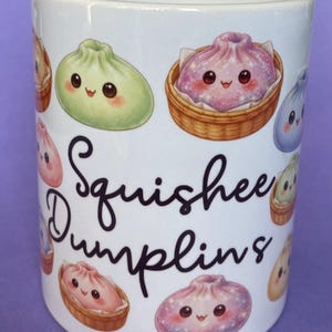 May include: White ceramic mug decorated with cartoon dumplings in different colours and baskets. The mug features the text "Squishie Dumplings" in a playful font. The dumplings have smiling faces.