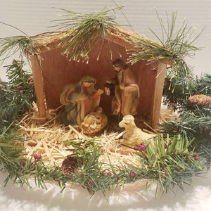 May include: A vintage wooden nativity scene with a manger, three figures, and a lamb. The scene is surrounded by a wreath of green pine branches with pine cones and red berries.