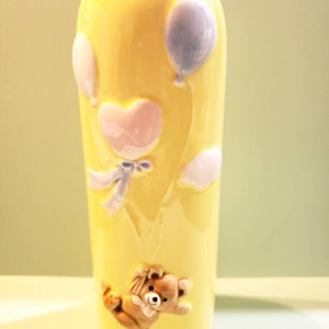 May include: A yellow ceramic vase with a teddy bear, heart, and balloons in relief. The vase has a green base.