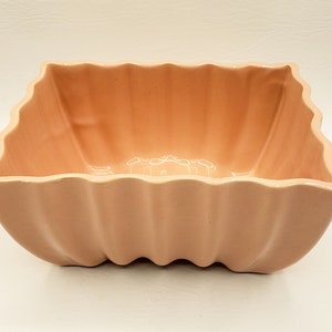 May include: A pale pink ceramic planter with a square shape and wavy edges. The planter has a smooth, glossy finish.