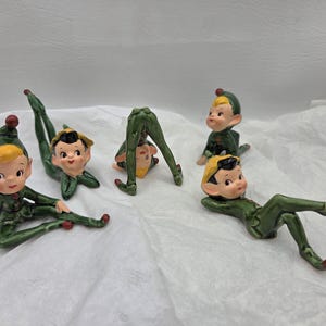 May include: A collection of ceramic elf figurines in various poses. The elves wear green outfits with yellow hair and red accents. The figurines are on a white surface.
