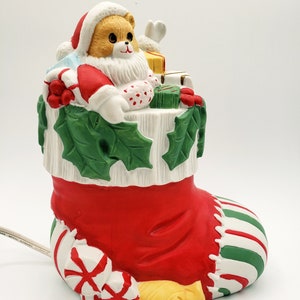 May include: A ceramic Christmas stocking with a Santa bear holding presents and a candy cane. The stocking is red, green, and white with a white base and a green holly design.